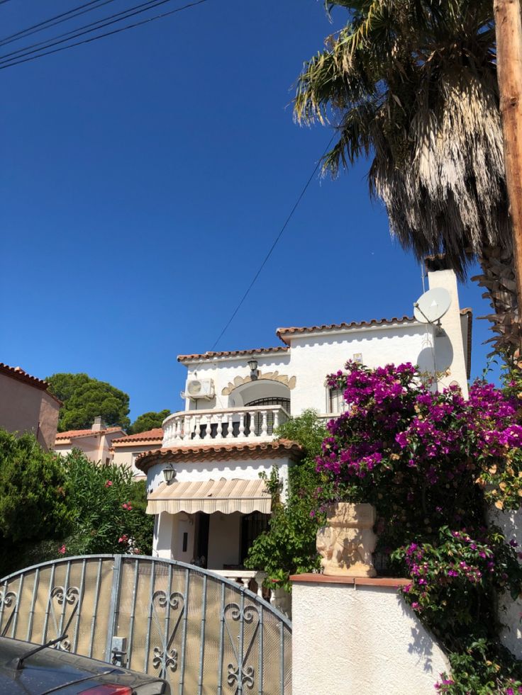 a white house with purple flowers and palm trees on the side of the road in front of it