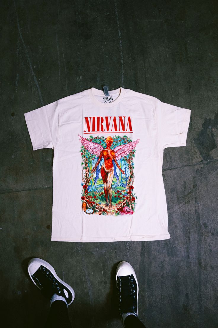 a white nirvana t - shirt sitting on top of a floor next to a pair of black shoes