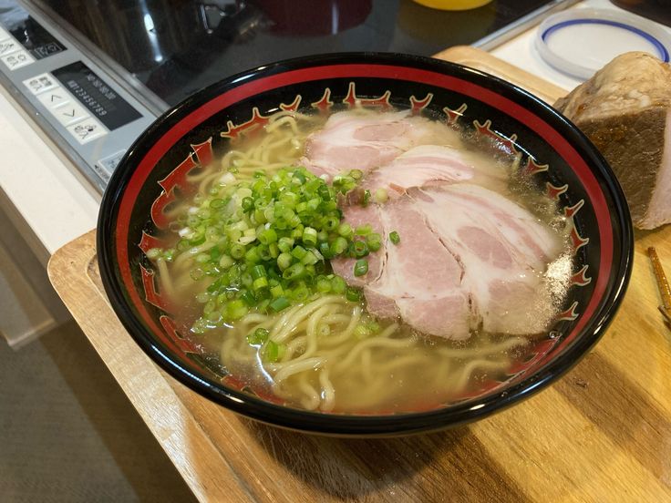 a bowl of ramen with meat and vegetables on a cutting board next to bread