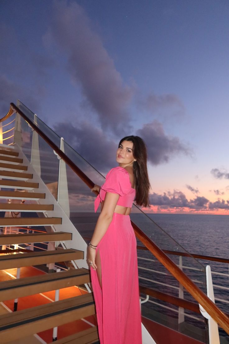 a woman in a pink dress standing on the deck of a cruise ship at sunset