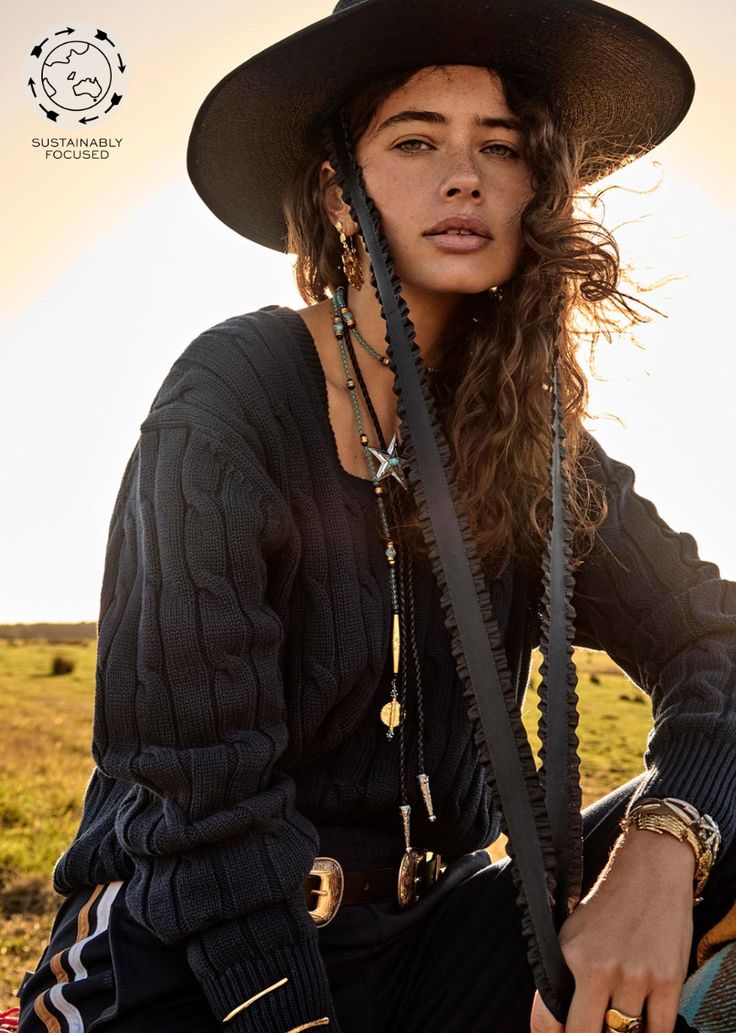 A woman with long, wavy hair wears a wide-brimmed black hat and a dark SEYMOUR KNIT SWEATER from The Upside, featuring a V-shaped neckline. She is adorned with various necklaces and gazes directly at the camera. The background showcases a grassy field bathed in sunset light, while the top left corner displays a circular logo with Sustainably Focused. Studio Images, Cotton Cable Knit Sweater, Arrow Logo, Back Neck, Beauty Body, Jeans Size Chart, Cable Knit Sweater, Leisure Wear, V Shape