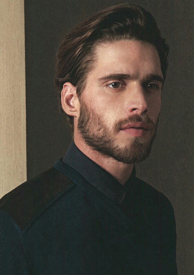 a man with long hair and beard standing in front of a wall wearing a black sweater