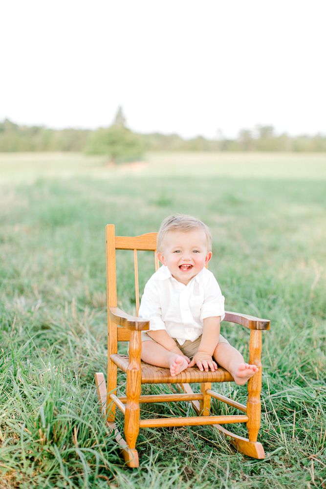 a toddler sitting in a wooden rocking chair on the grass smiling at the camera