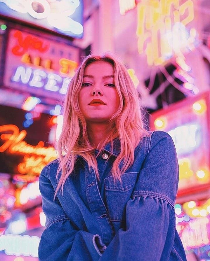 a blonde woman standing in front of neon signs and buildings with her hands on her hips