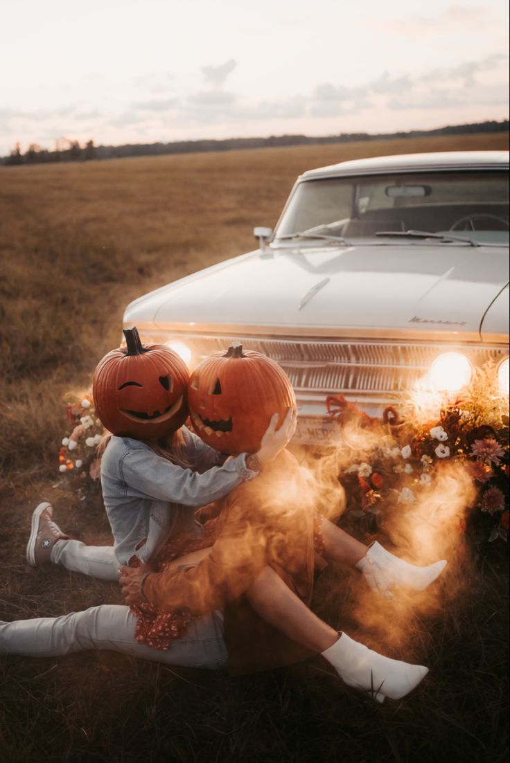 two people sitting in the grass with pumpkins on their heads and an old car behind them