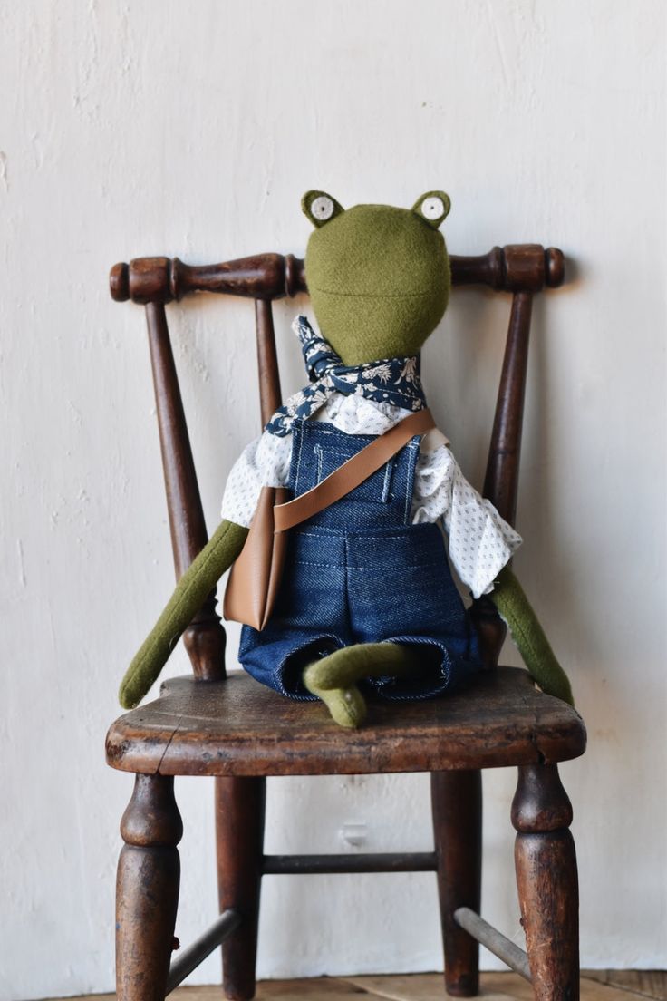 a stuffed frog sitting on top of a wooden chair with blue overalls and a bow tie