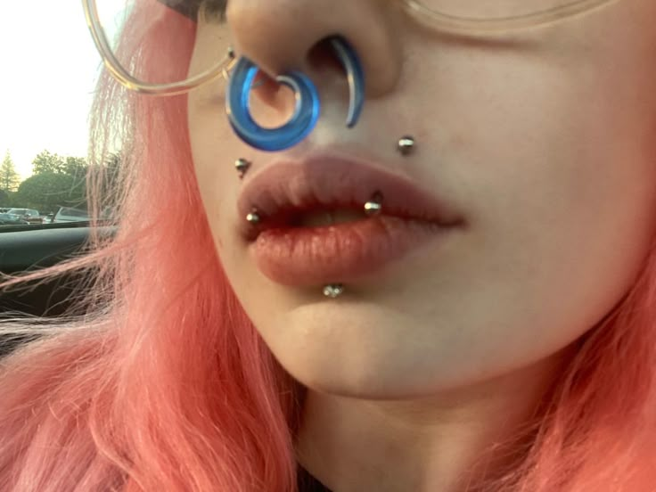 a close up of a person with pink hair and piercings on her nose wearing glasses