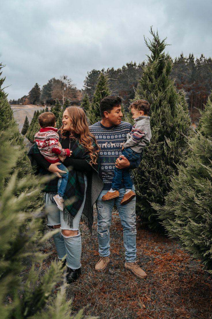 a man and woman holding two children in their arms while standing among christmas tree trees