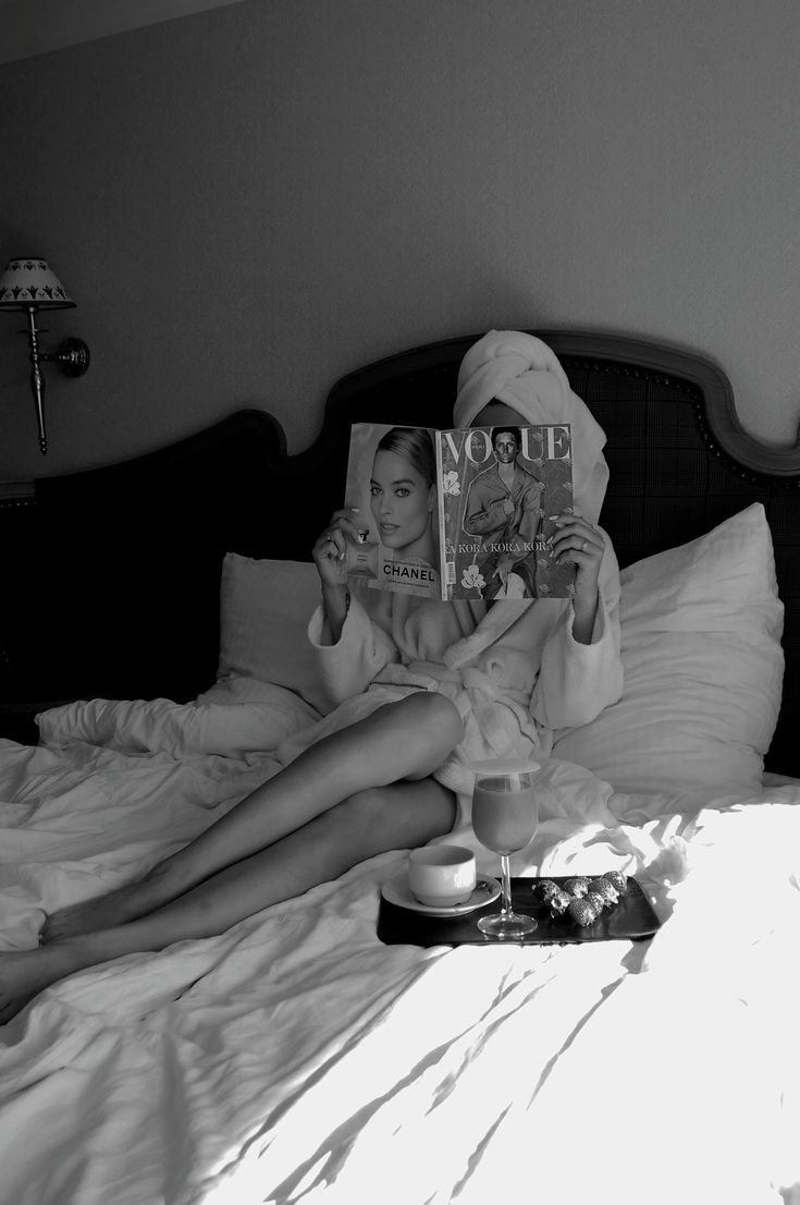 a woman is laying in bed reading a magazine and drinking coffee with her legs crossed