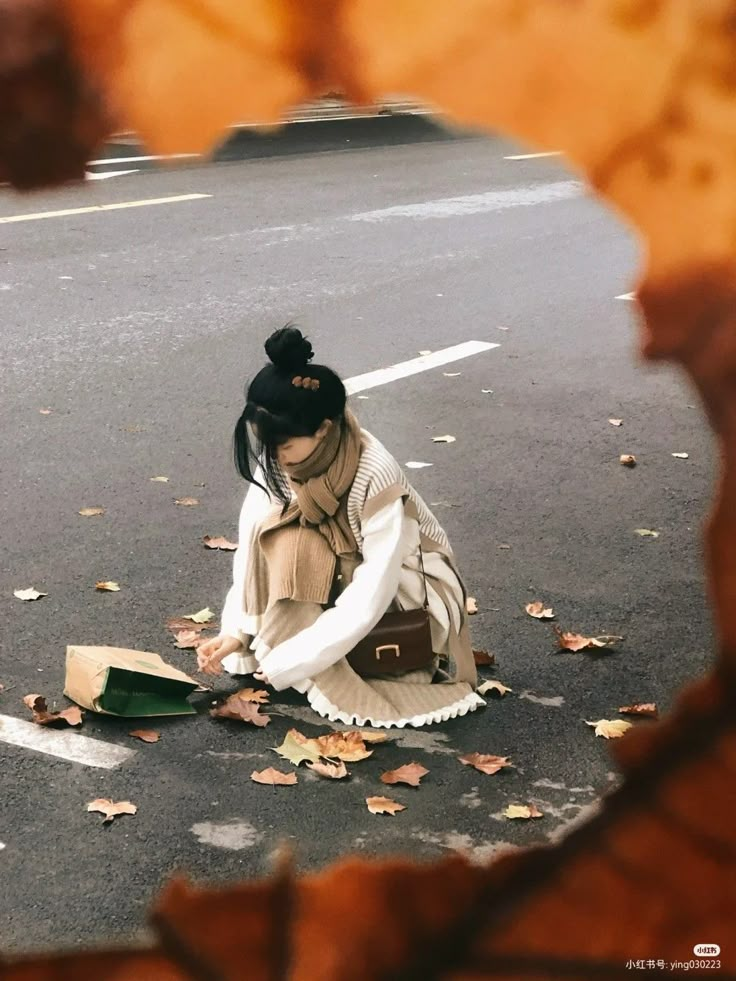 a woman kneeling down on the side of a road next to leaves and a book