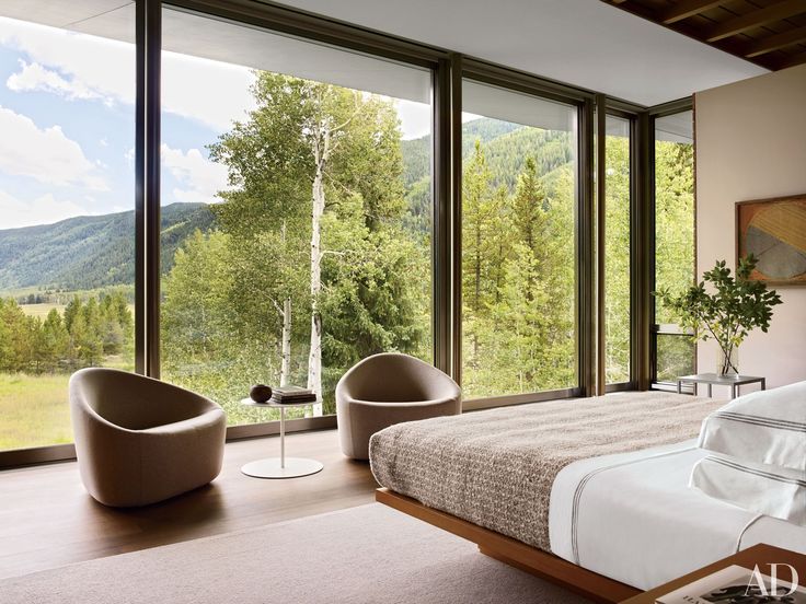 a bedroom with large windows overlooking the mountains and trees is pictured in this image, there are two chairs on either side of the bed