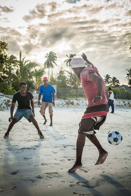 three men playing soccer on the beach with palm trees in the backgrouds