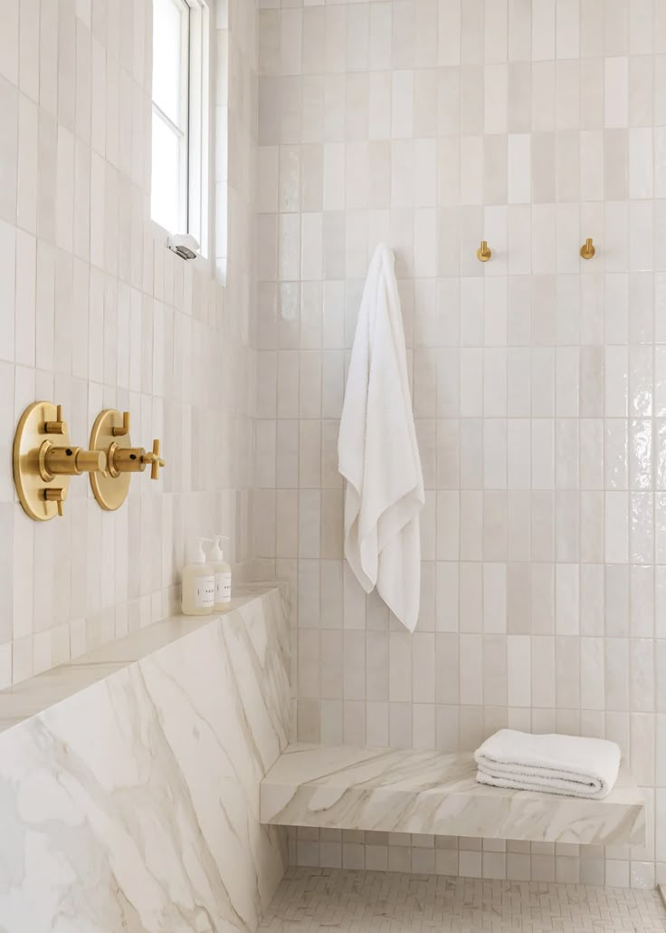 a white tiled bathroom with gold faucet and marble bench in the shower area