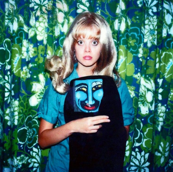 a woman with blonde hair and blue eyes holding a black bag in front of a green curtain