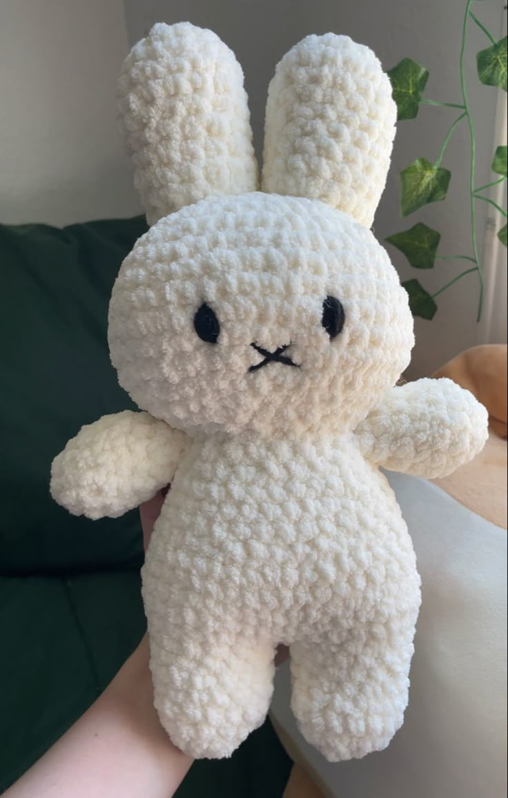 a hand holding a white crocheted stuffed animal with black eyes and ears, in front of a plant