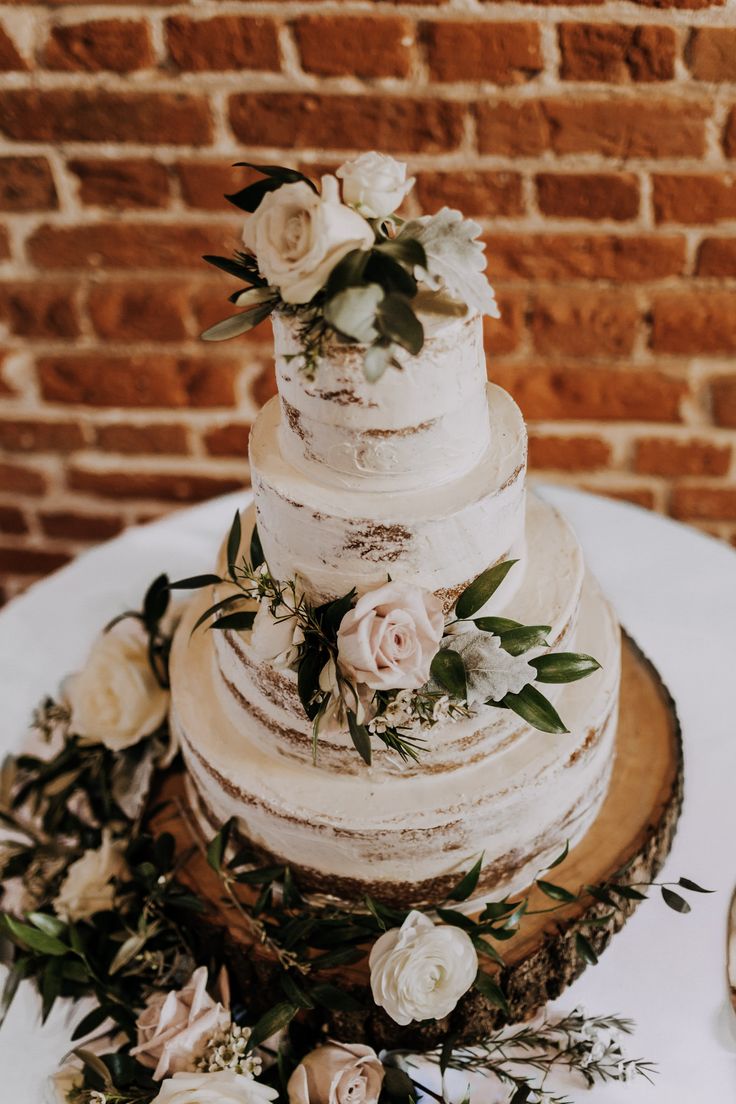 a wedding cake with white flowers and greenery sits on a table in front of a brick wall