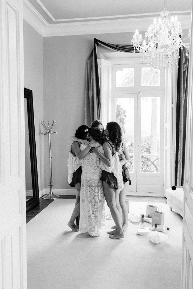 three girls hugging each other in front of a window with a chandelier hanging from the ceiling