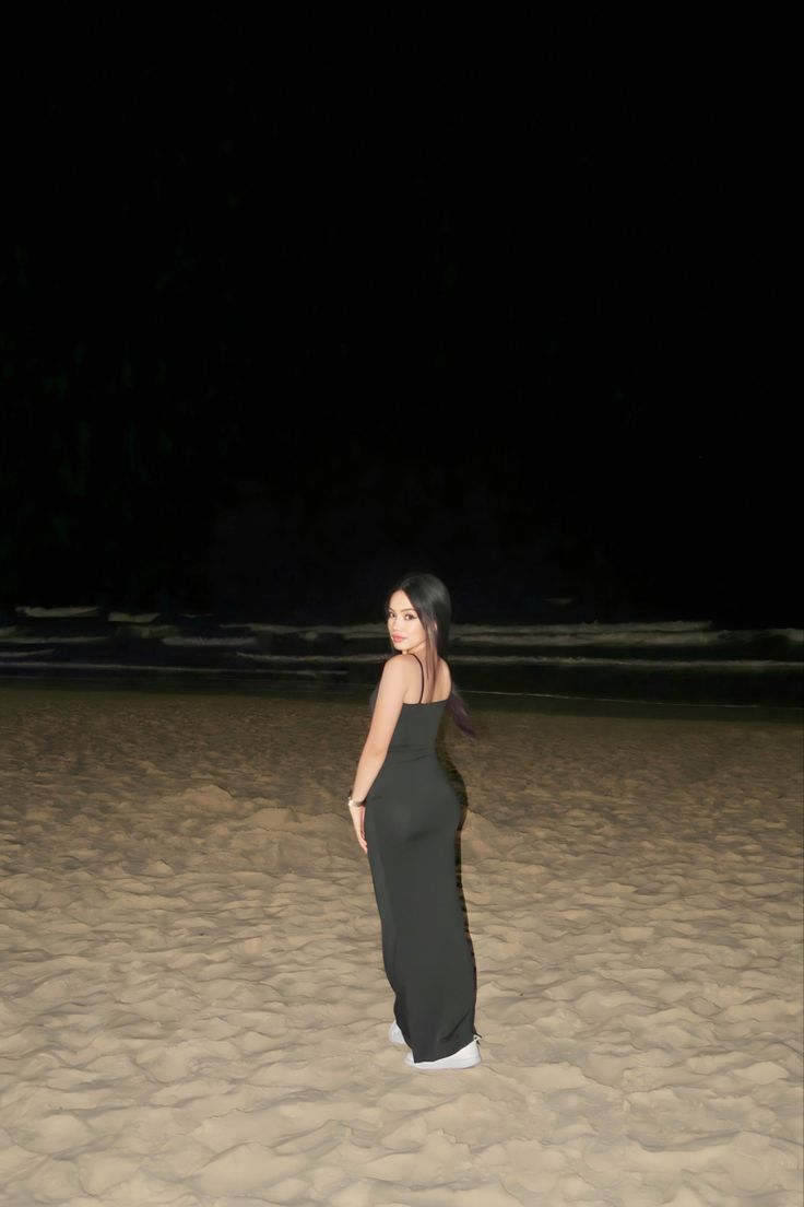 a woman standing on top of a sandy beach next to the ocean at night with her hands in her pockets