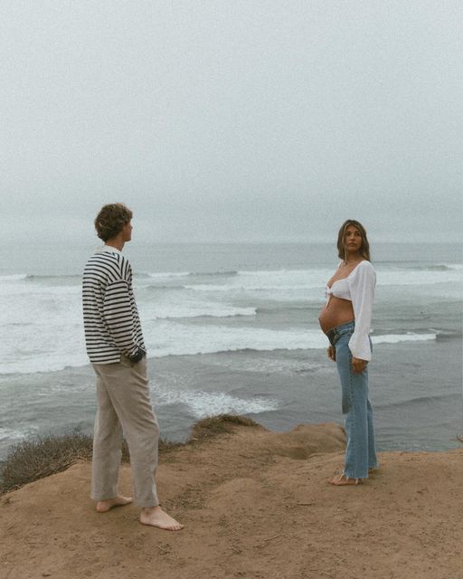 a pregnant woman standing on top of a cliff next to the ocean with her husband