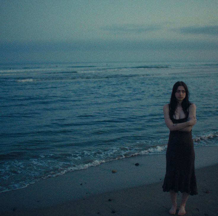 a woman standing on top of a sandy beach next to the ocean at night with her arms crossed