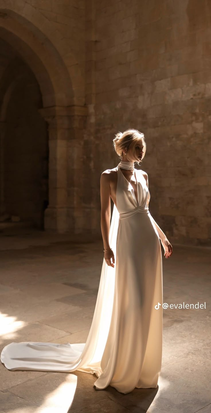 a woman in a white dress is standing near a stone wall and looking off into the distance