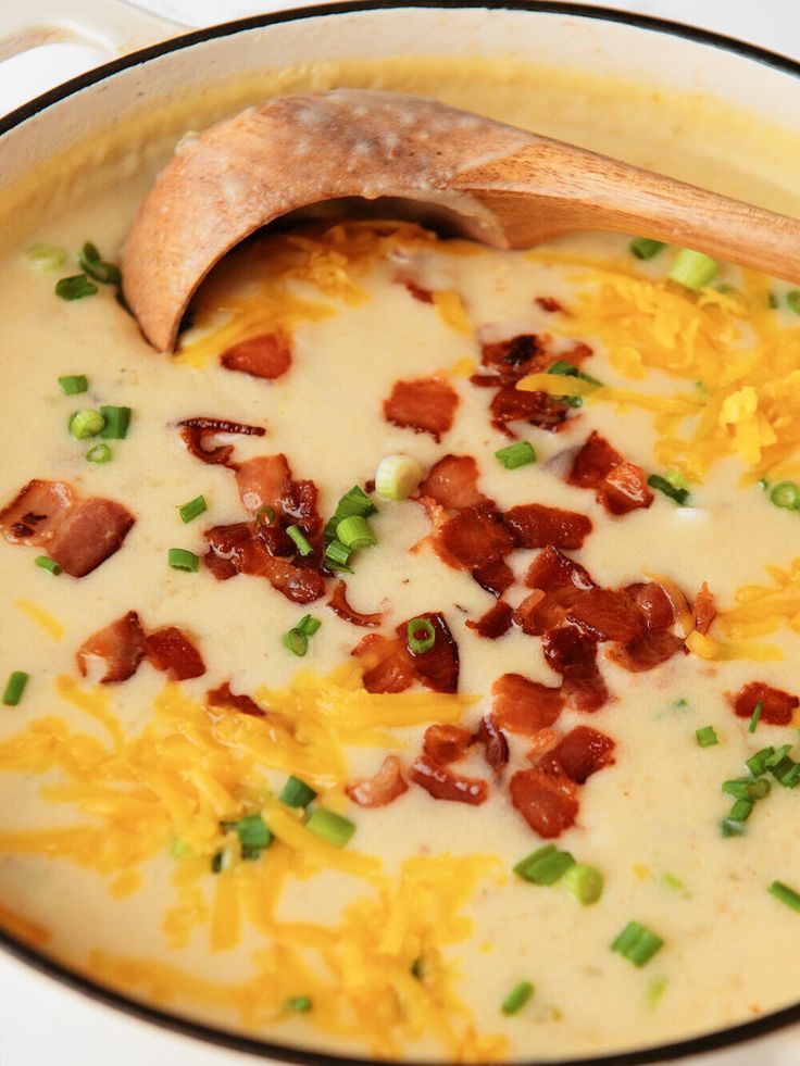 a bowl of soup with bacon, cheese and green onions in it next to a wooden spoon