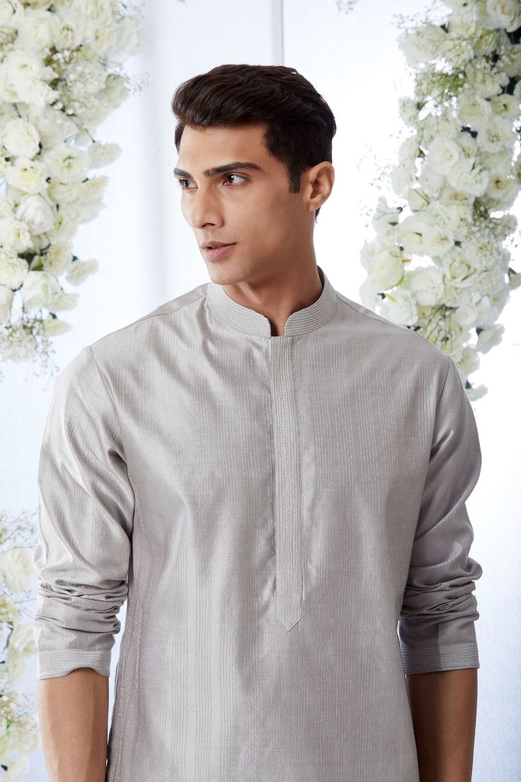 a man standing in front of white flowers wearing a grey shirt and pants with buttons on the chest