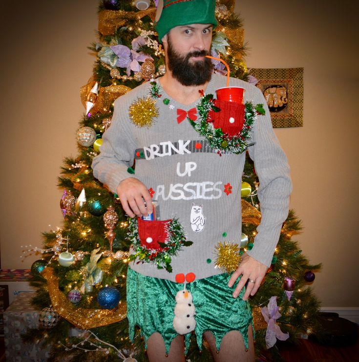 a man with a beard wearing green shorts and a christmas sweater standing in front of a christmas tree