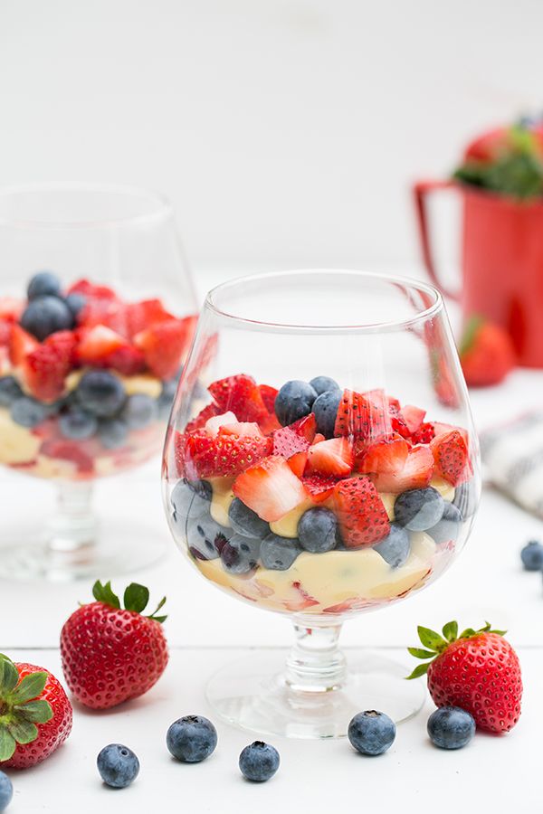 two glasses filled with fruit salad on top of a white table next to strawberries and blueberries