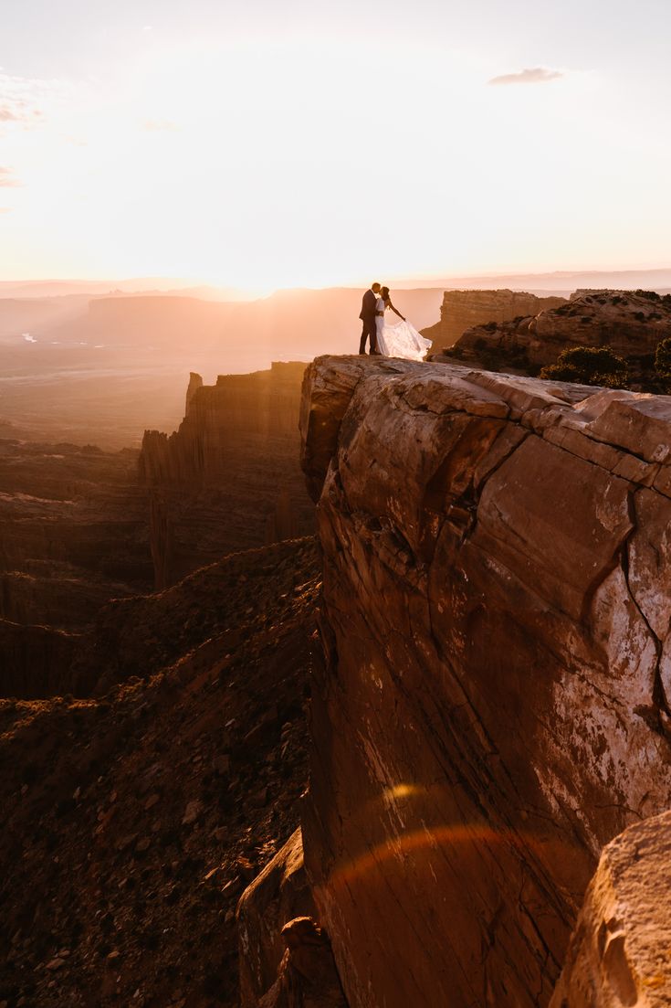 a man and woman standing on top of a rock formation with the sun setting in the background