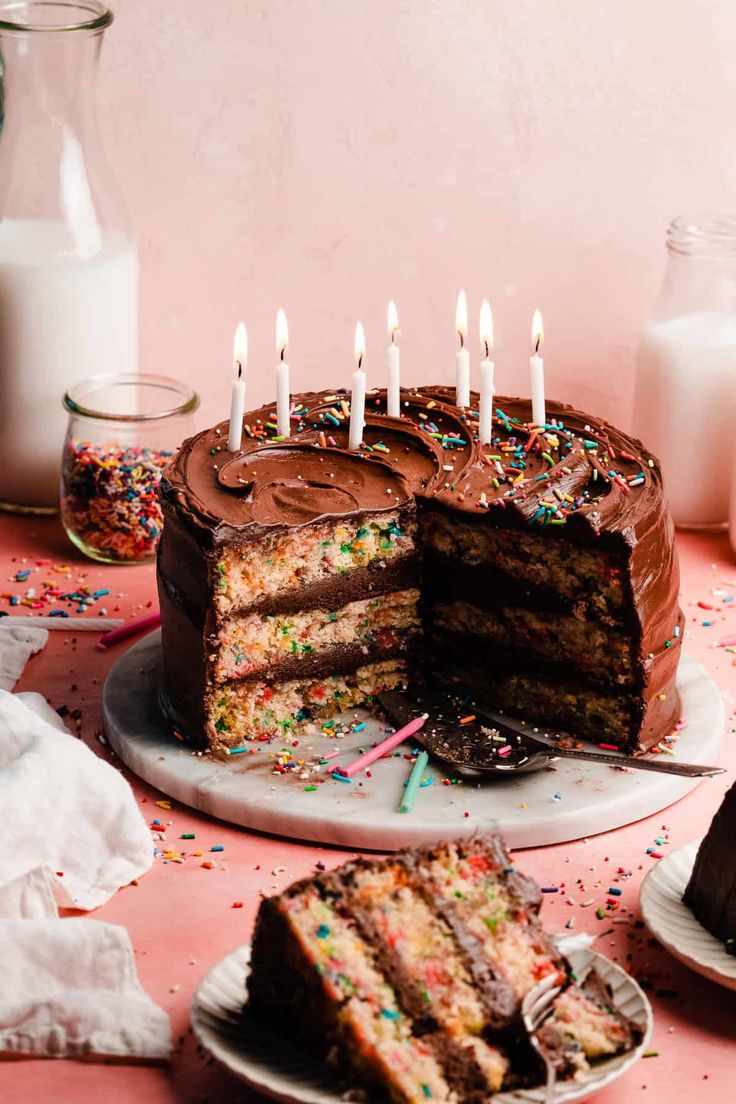 a birthday cake with lit candles is on a table next to other cakes and confetti