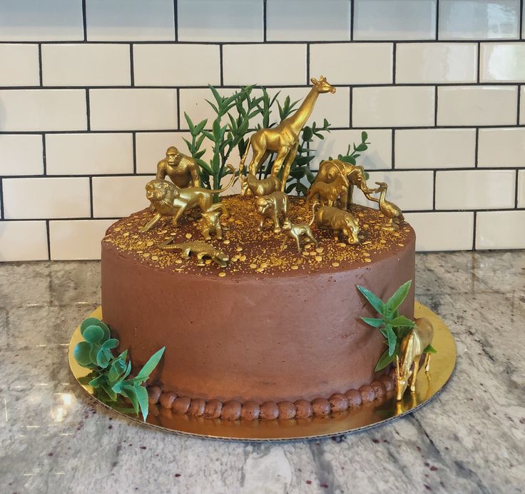 a chocolate cake decorated with gold animals and greenery on a marble countertop in front of a white brick wall