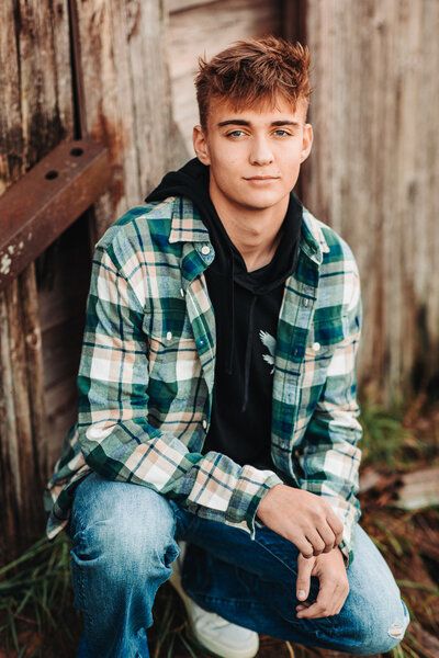 a young man sitting on the ground in front of a wooden fence wearing jeans and a flannel shirt