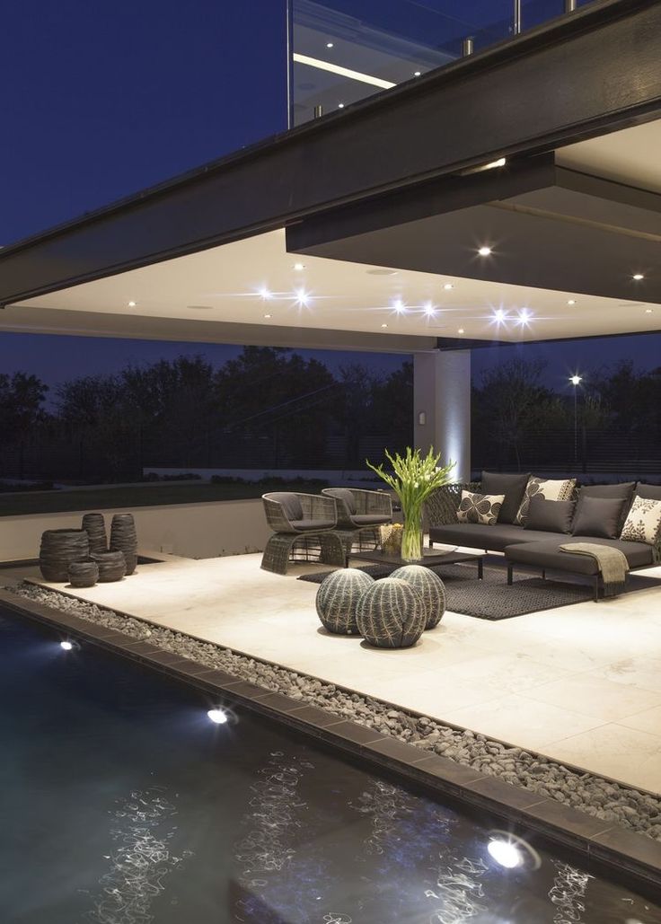 an outdoor living area with couches and tables next to a swimming pool at night