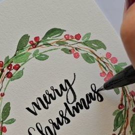 someone is writing merry christmas on a card with a pen and watercolor pencils