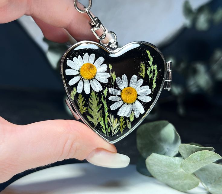 a heart shaped keychain with daisies painted on the front and sides, held by a person's hand