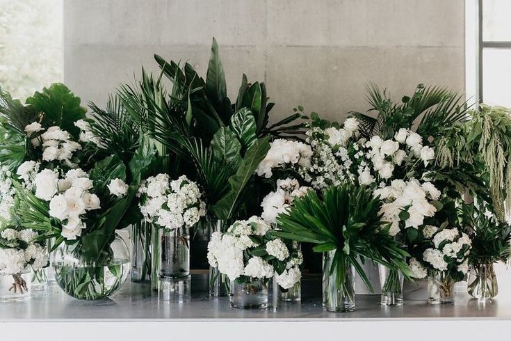 white flowers and greenery are arranged in vases on a table with concrete walls