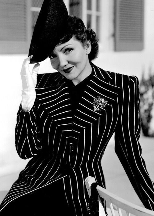 a woman in a pinstripe suit and hat sitting on a chair with her hand up to her face