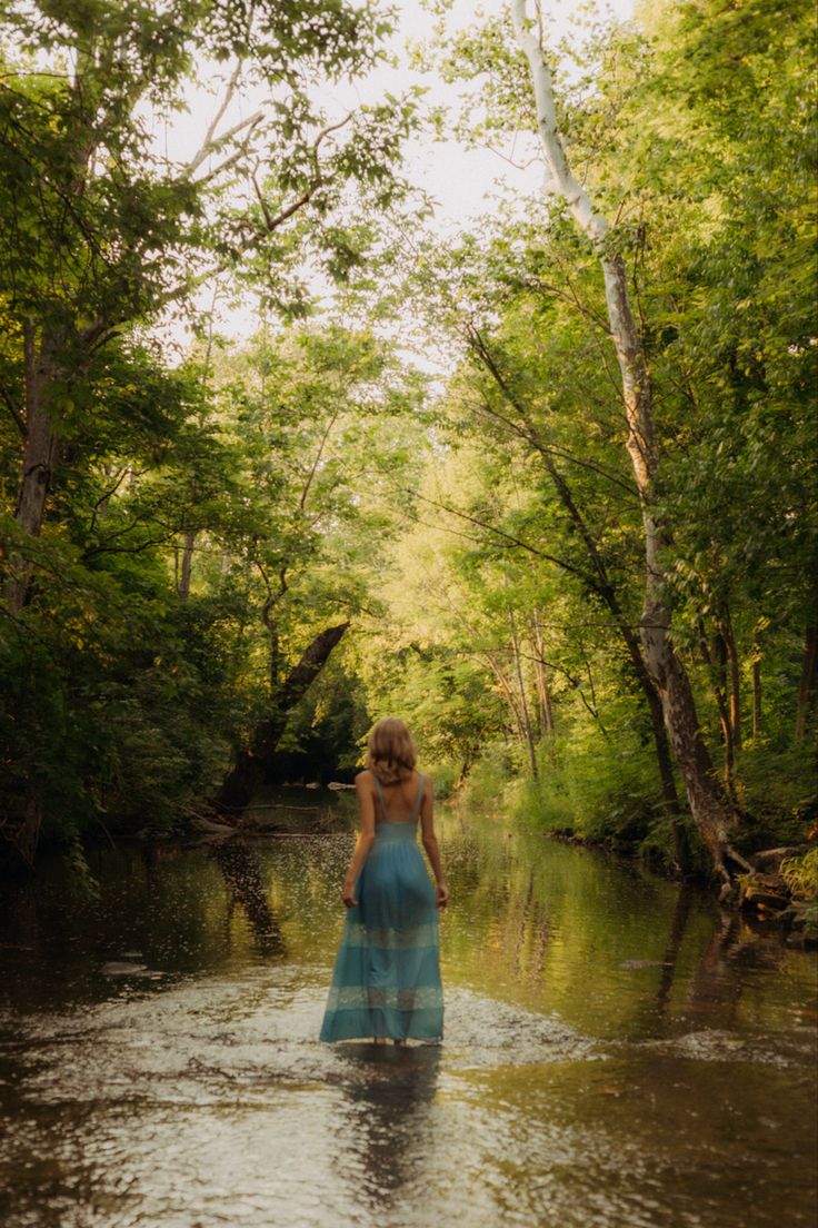 a woman in a blue dress standing in the middle of a river with trees around her