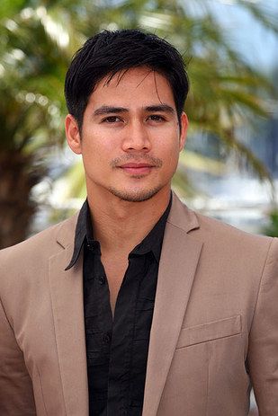 a man in a tan suit and black shirt posing for the camera with palm trees behind him