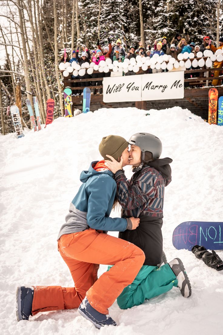 two snowboarders kissing in the snow near a ski slope with many people watching