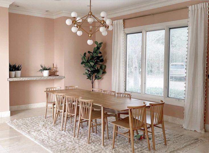 a dining room with pink walls and white curtains on the window sill, wooden table surrounded by chairs