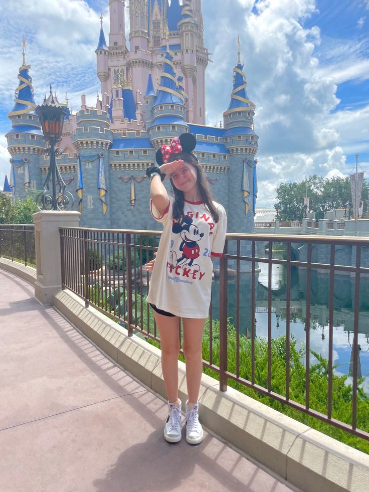 a woman standing in front of a castle with a mickey mouse t - shirt on