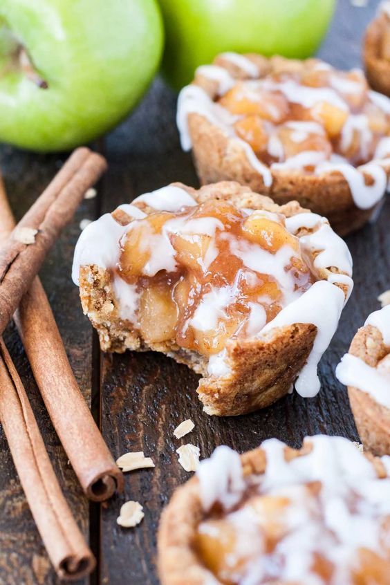 apple pies with white icing and cinnamon sticks on a wooden table next to apples