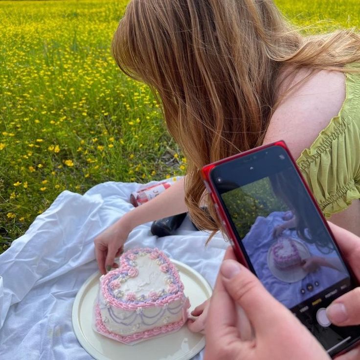 a woman taking a photo of a cake on her phone while sitting in the grass