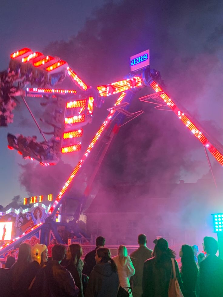 people are standing around in front of a ferris wheel at an amusement park during the night