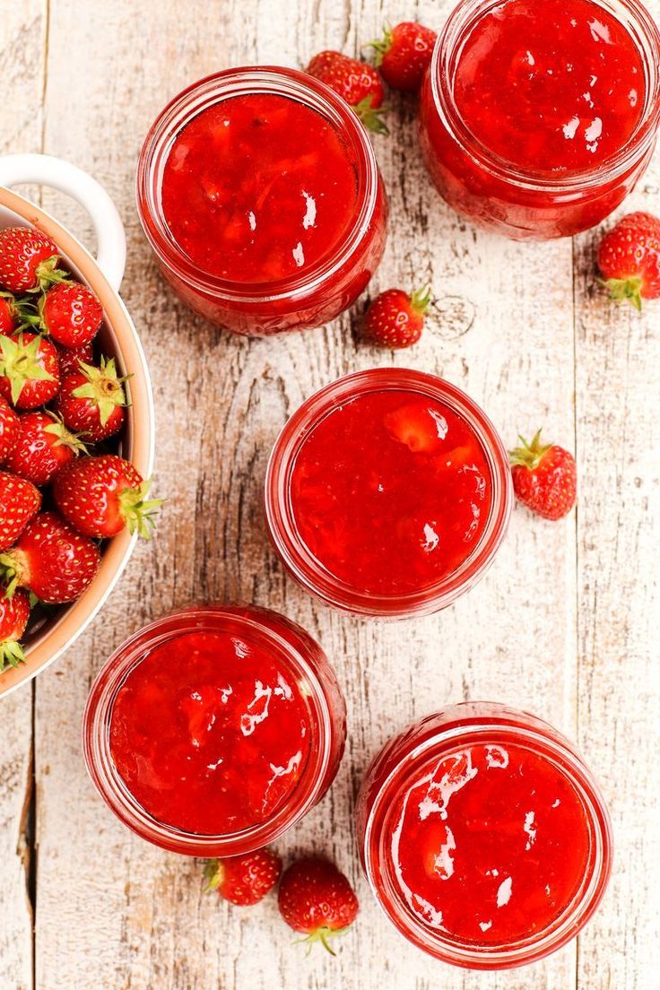 four jars filled with strawberry jam on top of a white wooden table next to strawberries