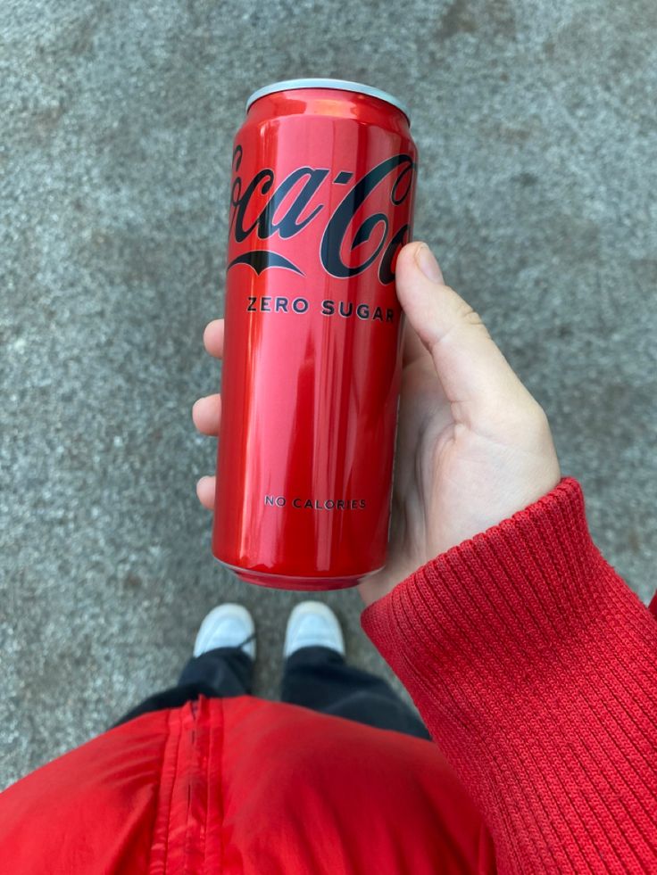 a person holding up a red can of coca - cola in their hand on the ground