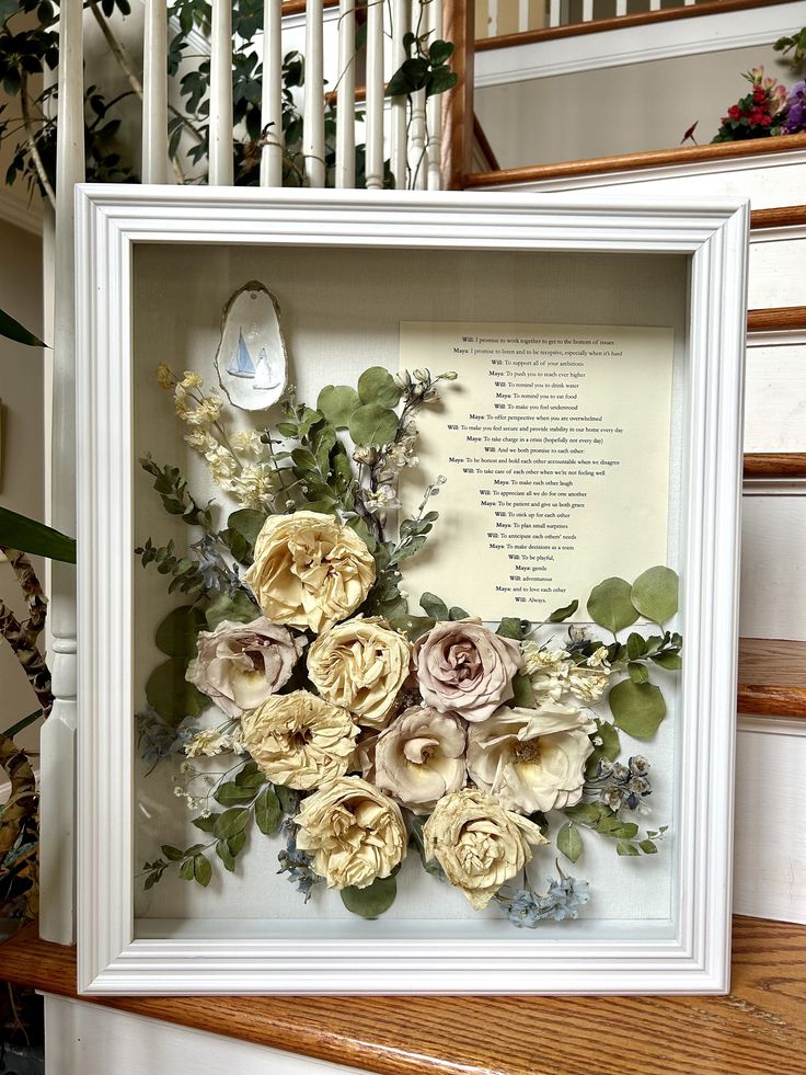 a white frame with some flowers in it on top of a wooden table next to stairs