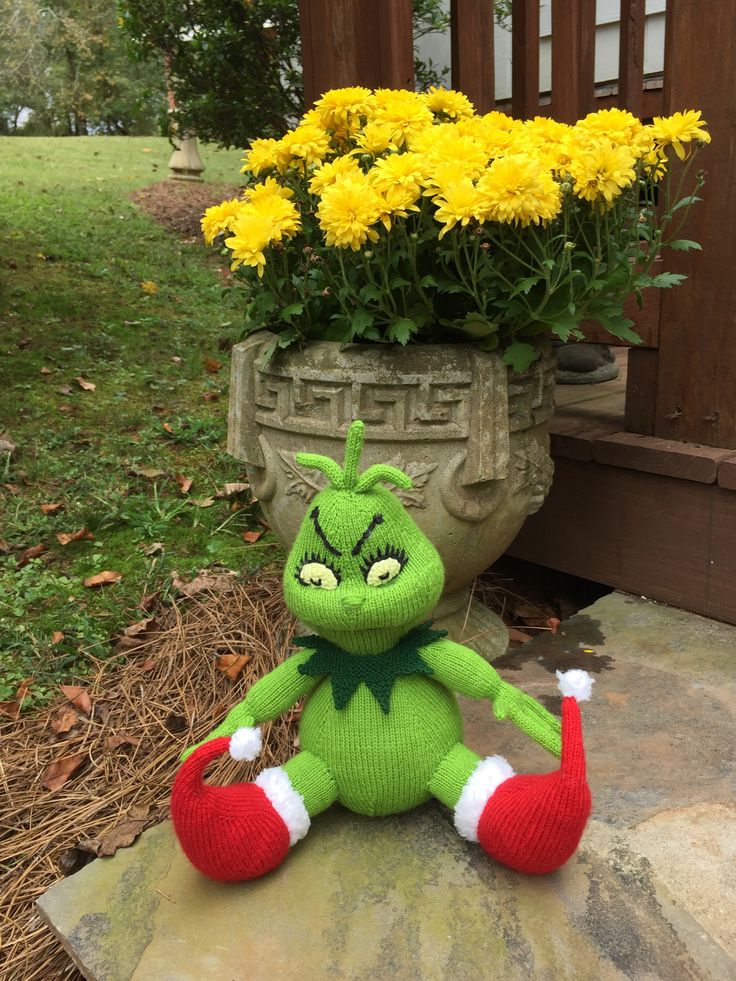 a green stuffed toy sitting on top of a stone slab next to yellow flowers in a pot